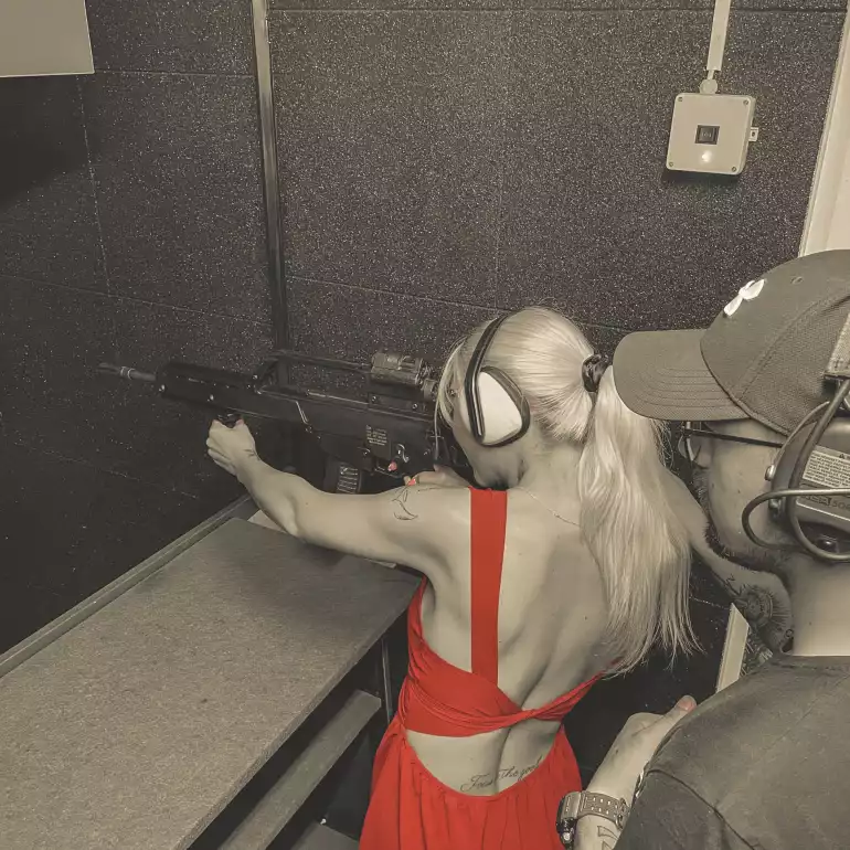 Blonde girl in red dress at a gun range in Budapest shooting a pistol under a supervision of an instructor.