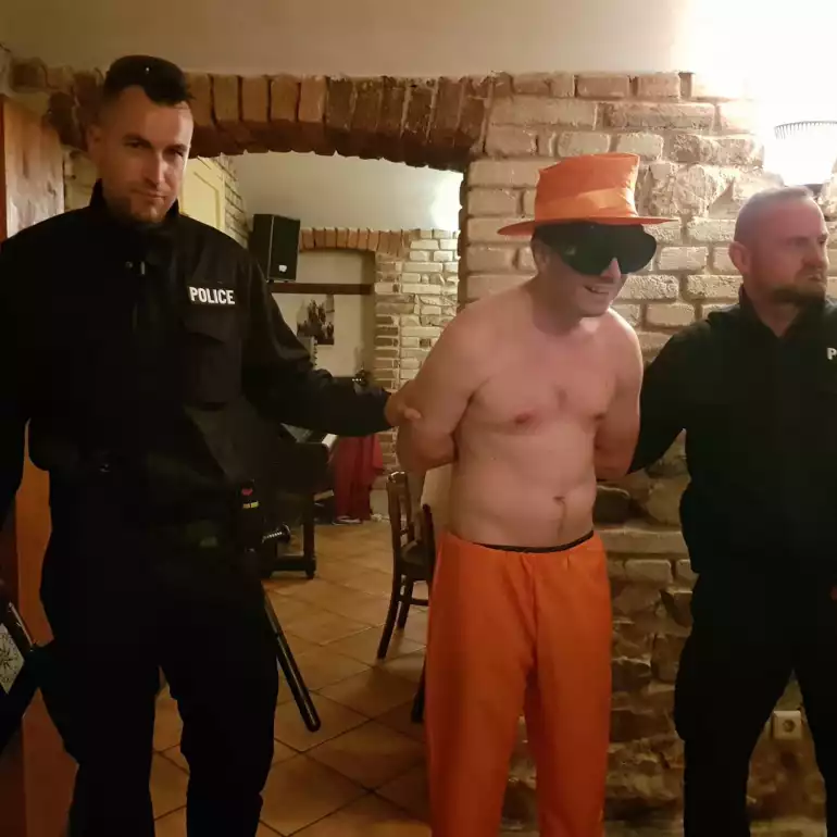Two man in black uniforms standing next to a handcuffed guy dressed all in orange with a hat.